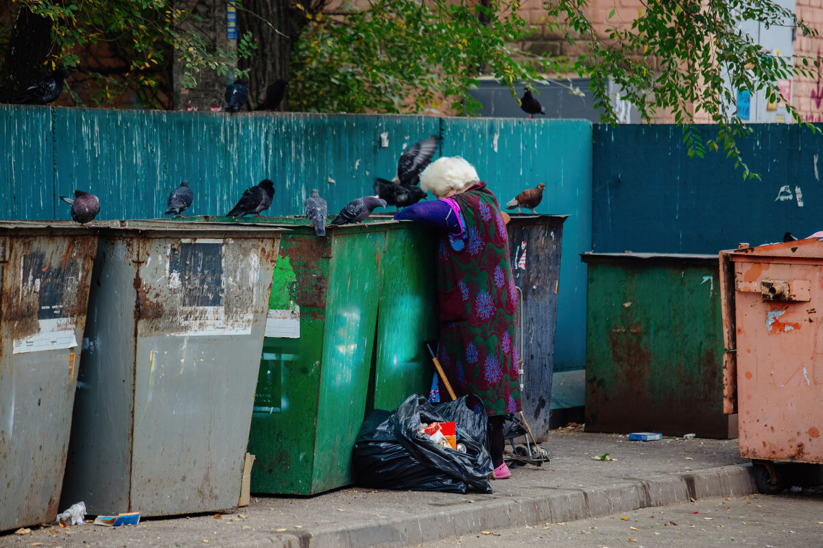 Homeless old woman looking into trash box in search for food. Homelessness problem concept